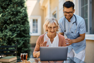 Happy senior woman making video call over laptop with help of male nurse on patio.