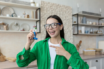 Young beautiful woman in glasses and green shirt at home holds asthma and allergy inhaler in hand,...