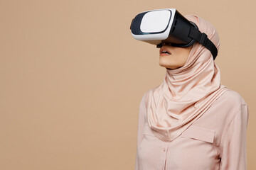 Young arabian asian muslim woman she wearing abaya hijab pink clothes watch in vr headset pc gadget isolated on plain pastel light beige background People uae middle eastern islam religious concept.