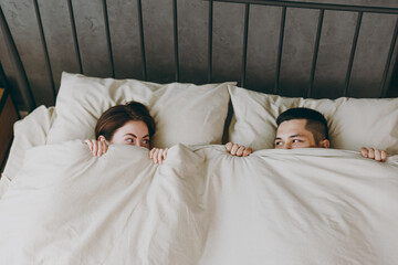Young couple two family man woman she he wear t-shirt pajama lying in bed cover face mouth with blanket look to each other spend time together in bedroom home in own room hotel. Real estate concept.