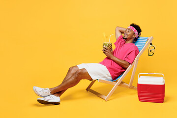 Full body young man he wear pink t-shirt bandana lying on deckchair near hotel pool drink pineapple juice hold hand behind neck isolated on plain yellow background. Summer vacation sea rest concept.