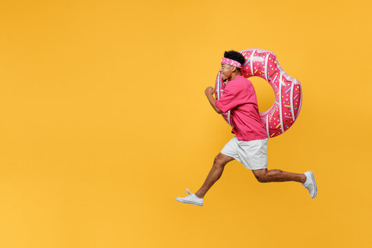 Side view happy young man he wear pink t-shirt near hotel pool jump high run fast hurrying up hold inflatable donut rubber ring isolated on plain yellow background. Summer vacation sea rest concept.