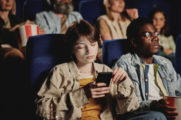 Young gen Z Caucasian woman getting bored with movie scrolling news feed in social network on smartphone at cinema