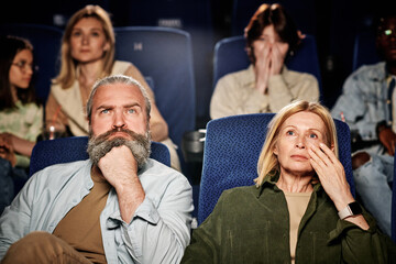 Selective focus of mature Caucasian man and woman watching drama movie at cinema, woman wiping...