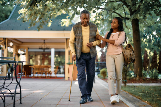 Happy African American woman walks with her senior father while visiting him at nursing home.