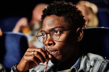 Close-up of young gen Z Black man wearing eyeglasses sipping fizzy drink while watching movie at...