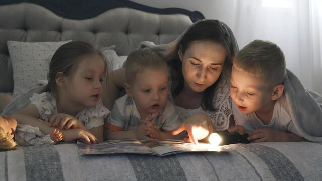 Mom in the evening with a flashlight reads a book to children covering them with a blanket. Close-up.
