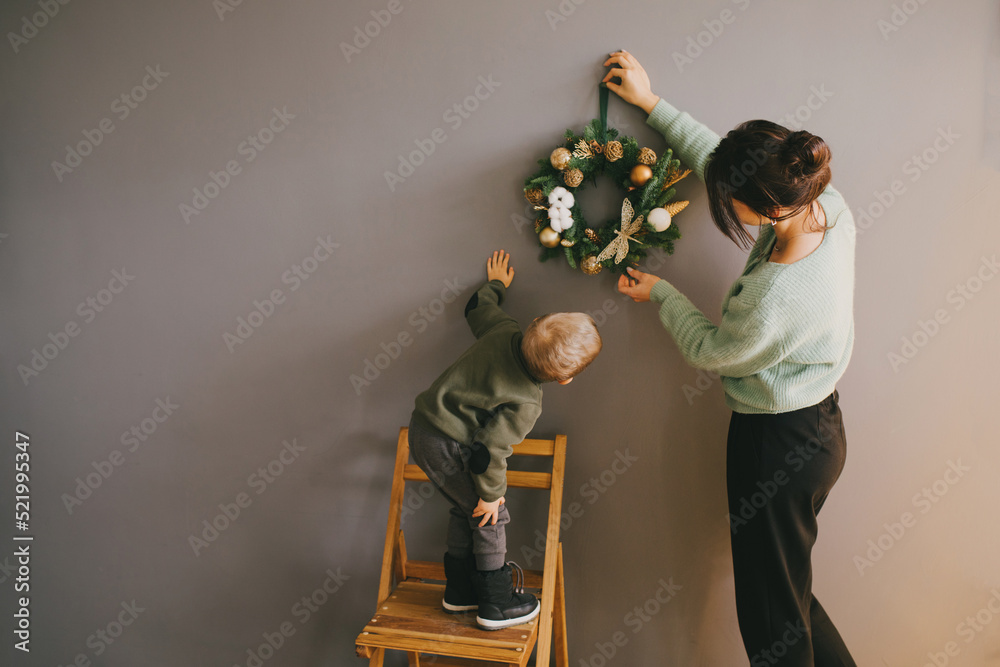 Wall mural mother and her little son decorating grey wall with handmade christmas wreath. festive home decorati