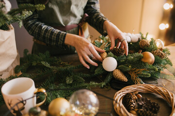 Millennial woman making Christmas wreath using pine branches and festive decorations in workshop....