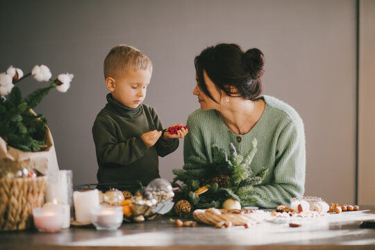 Mother and her little son making Christmas wreath together using pine branches and cones. Festive holiday cozy home decorations.