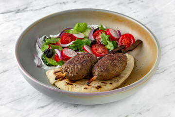 Lula kebab on skewers, pita bread and fresh vegetable salad in a  plate on a marble background. Restaurant banquet menu.