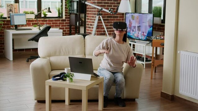 Young adult person wearing futuristic virtual reality headset enjoying cyberspace while sitting on sofa at home. Playful woman with VR goggles playing on metaverse inside living room.