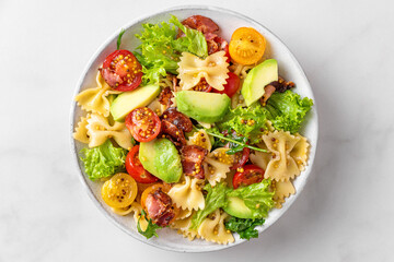 Pasta farfalle salad with tomato, bacon, avocado and mustard dressing in a plate on white table....