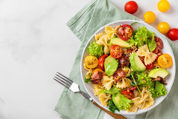 Cold summer pasta salad with bacon, tomatoes, avocado and mustard in a plate with fork on white background. Top view