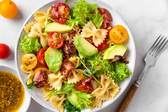 Delicious italian salad made of pasta farfalle, tomatoes, avocado, bacon and mustard in a plate on white table. Top view
