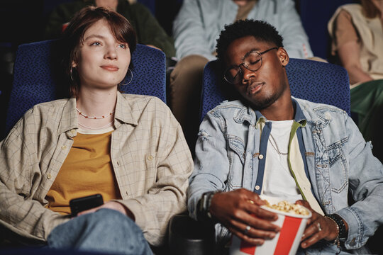 Horizontal medium portrait of young Black man having nap while his Caucasian girlfriend watching movie at cinema