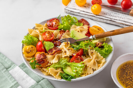 Cold summer pasta salad with bacon, tomatoes, avocado and mustard in a plate with fork on white background