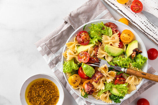 Delicious italian salad made of pasta farfalle, tomatoes, avocado, bacon and mustard in a plate with fork on white table