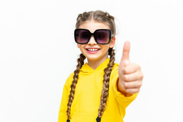 A teenager gives a thumbs up on a white isolated background. Portrait of a little girl in a yellow tracksuit and sunglasses.