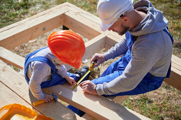 Father with toddler son building wooden frame house. Male builder and kid playing with tape measure...