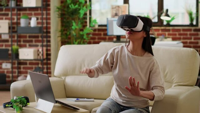 Playful woman wearing virtual reality headset scrolling through metaverse cyberspace while working from home. Asian person wearing futuristic VR goggles while interacting with cyber reality.
