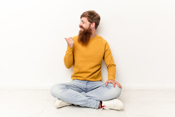 Young caucasian man sitting on the floor isolated on white background points with thumb finger away, laughing and carefree.
