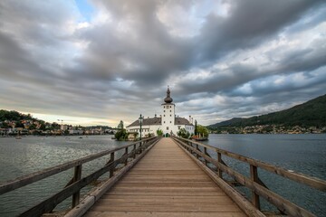 Obraz premium Schloss Ort on Traunsee lake near Gmunden, Austria. The view on the wooden pier leading towards the church. Cloudy sky with sunset in the background. 