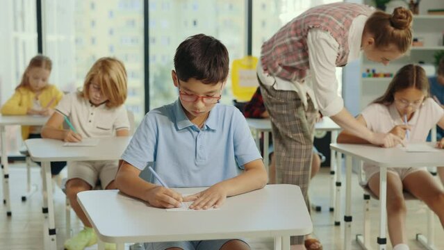 Asian boy pupil wearing glasses sits at a first desk in the classroom and fills out tests, and the teacher in the background help students with their test. People and educational concept.