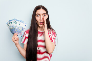Young caucasian woman holding banknotes isolated on blue background is saying a secret hot braking...