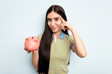 Young caucasian woman holding a piggy bank isolated on blue background pointing temple with finger, thinking, focused on a task.