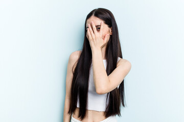 Fototapeta premium Young caucasian woman isolated on blue background blink at the camera through fingers, embarrassed covering face.