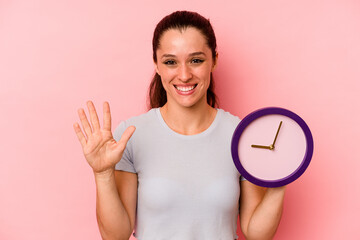 Young caucasian woman holding a clock isolated on pink background smiling cheerful showing number five with fingers.