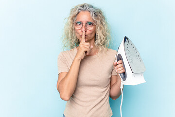 Middle age caucasian woman holding an iron isolated on blue background keeping a secret or asking for silence.