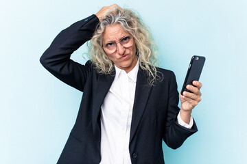 Middle age business caucasian woman holding mobile phone isolated on blue background being shocked,...