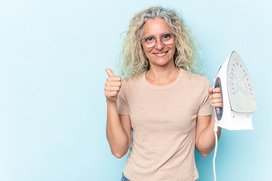 Middle age caucasian woman holding an iron isolated on blue background smiling and raising thumb up