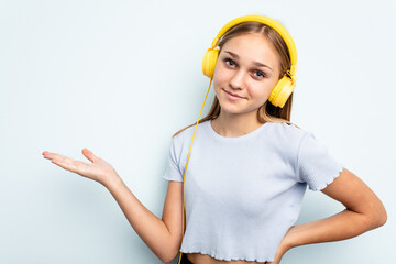 Young caucasian girl listening to music isolated on blue background showing a copy space on a palm...