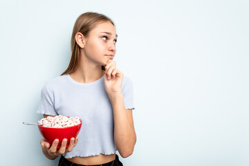 Young caucasian girl eating cereals isolated on blue background looking sideways with doubtful and skeptical expression.