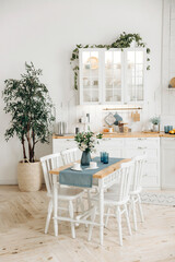 Wooden dining table with a bouquet of fresh flowers in a vase and white chairs in a modern white Scandinavian style kitchen with kitchen accessories and plants in pots. Empty space.