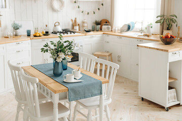 Wooden dining table with a bouquet of fresh flowers in a vase and white chairs on the blured background of modern white Scandinavian style kitchen with kitchen accessories and plants in pots. Nobody
