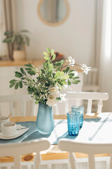 A bouquet of fresh flowers in a glass vase with sunbeams on a dining table with two blue glasses against a blurred background of a white Scandinavian style kitchen.