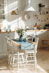 A wooden dining table with a bouquet of fresh flowers in a vase and white chairs in a modern white Scandinavian style kitchen with kitchen accessories and sunbeams on the wall and floor. Empty space.