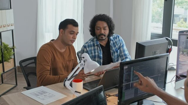 Medium slowmo of Indian team of programmers discussing documents and writing program codes while sitting by computers at desk in office
