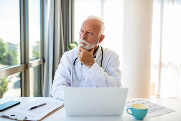 Serious mature doctor using laptop and sitting at desk. Senior professional medic physician wearing...