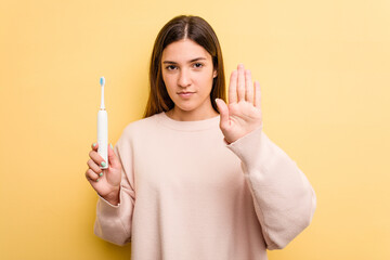 Young caucasian woman holding a electric toothbrush isolated on yellow background standing with...