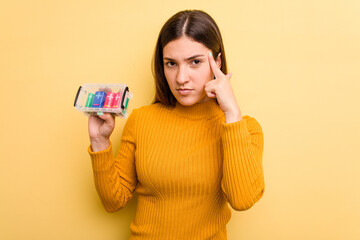 Young caucasian woman holding a batteries box isolated on yellow background pointing temple with finger, thinking, focused on a task.