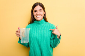 Young caucasian woman holding measuring jug isolated on yellow background person pointing by hand to a shirt copy space, proud and confident