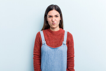 Young caucasian woman isolated on blue background blows cheeks, has tired expression. Facial...
