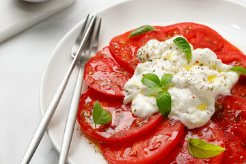 Salad with Stracciatella cheese on white plate with fork, oil, fresh basil and tomatoes. Italian Stretched curd cheese product in Stracciatella salad. Close up
