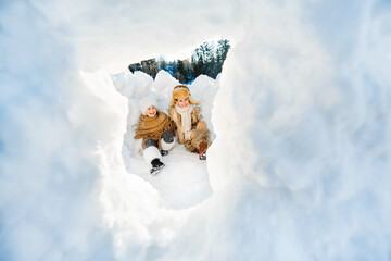 Happy family have fun playing in snow hut fort on sunny winter day. Children look out the window in snow wall.