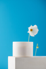 Mockup of a white jar with nourishing face and body cream on a pedestal on a blue background with a flower. Skin care.